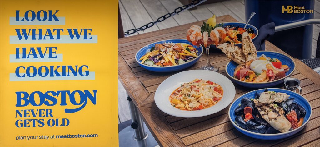 A colorful outdoor table displays five plates of seafood dishes next to a yellow sign that reads, “Look what we have cooking. Boston never gets old. Plan your stay at meetboston.com.”.