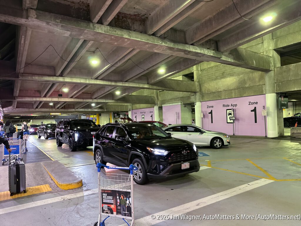 Cars line up in an indoor parking garage at a designated ride app pickup zone, with signage and a shopping cart visible in the foreground.