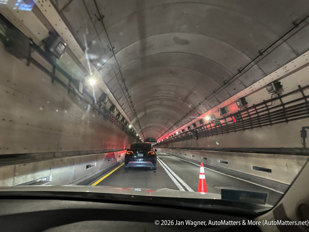 A view from inside a car driving through a tunnel, following other vehicles. Orange traffic cone and tunnel lighting are visible.