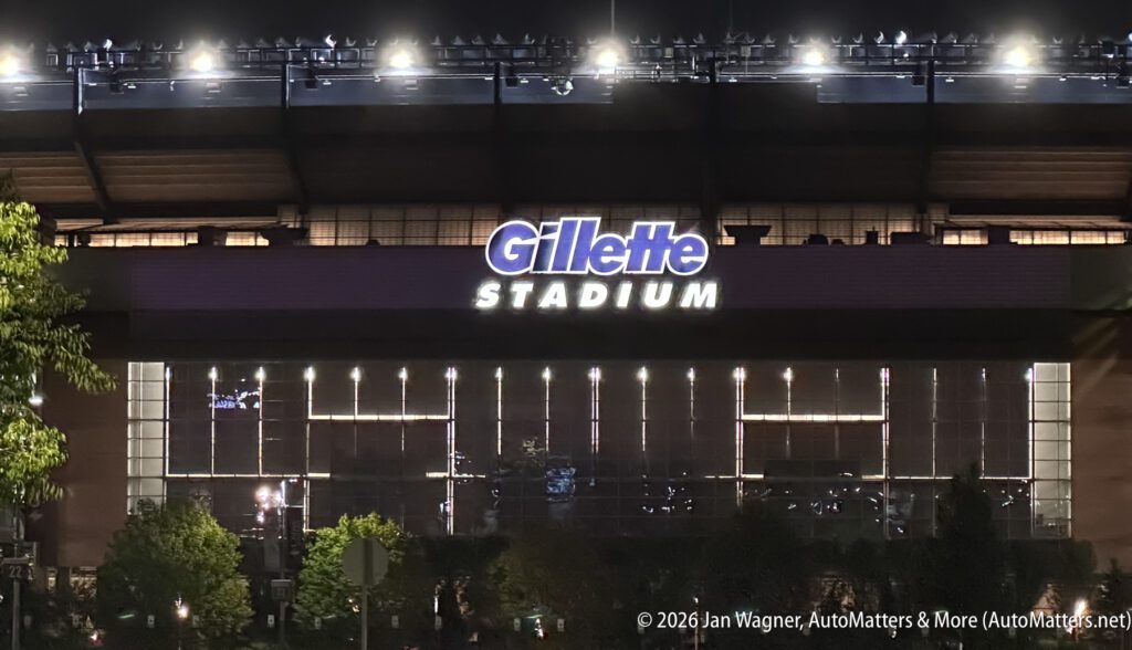Exterior nighttime view of Gillette Stadium, with its illuminated sign visible above glass windows and trees in the foreground.