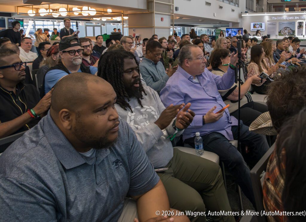 Audience members seated closely together, some clapping and others holding phones, at an indoor event with bright lighting and visible background signage.
