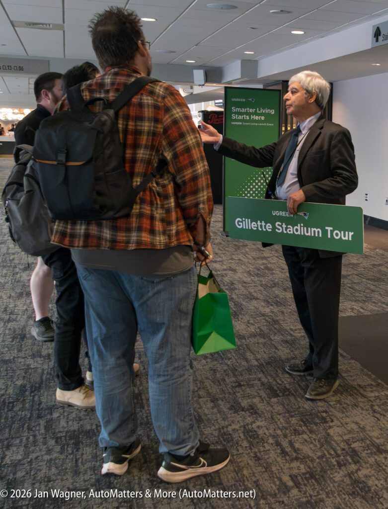 A man in a suit holds a “Gillette Stadium Tour” sign and talks to a small group of casually dressed people inside a building.