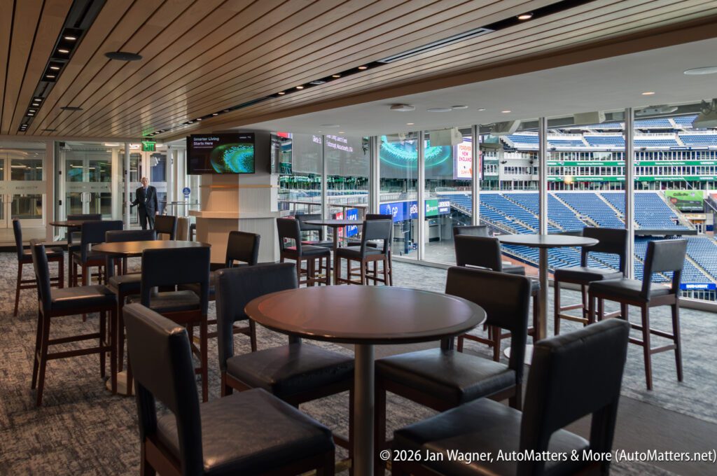 Modern stadium lounge with empty tables and chairs, large windows overlook seating and field, digital screens visible; one person stands by the window.
