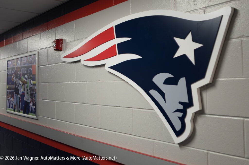 Large New England Patriots logo mounted on a gray hallway wall with a framed football photograph visible in the background.