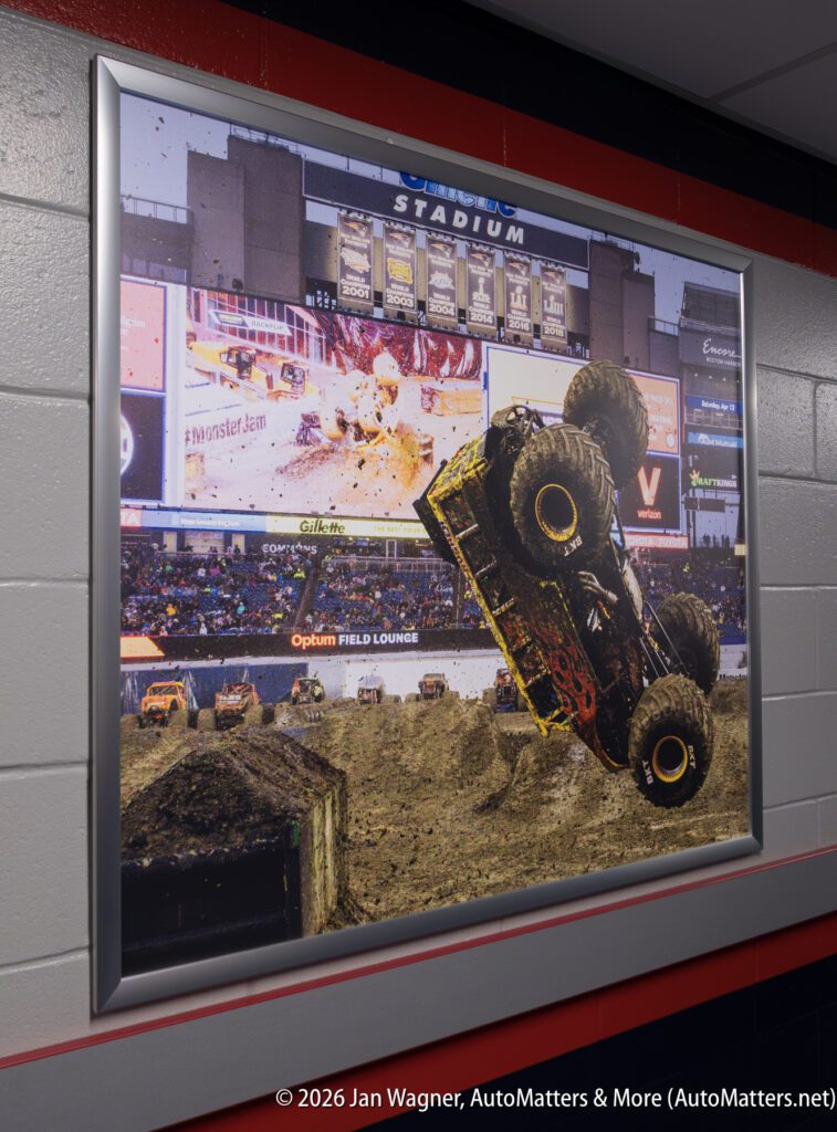 Framed photograph of a monster truck performing a wheelie at an indoor stadium event, mounted on a tiled wall.