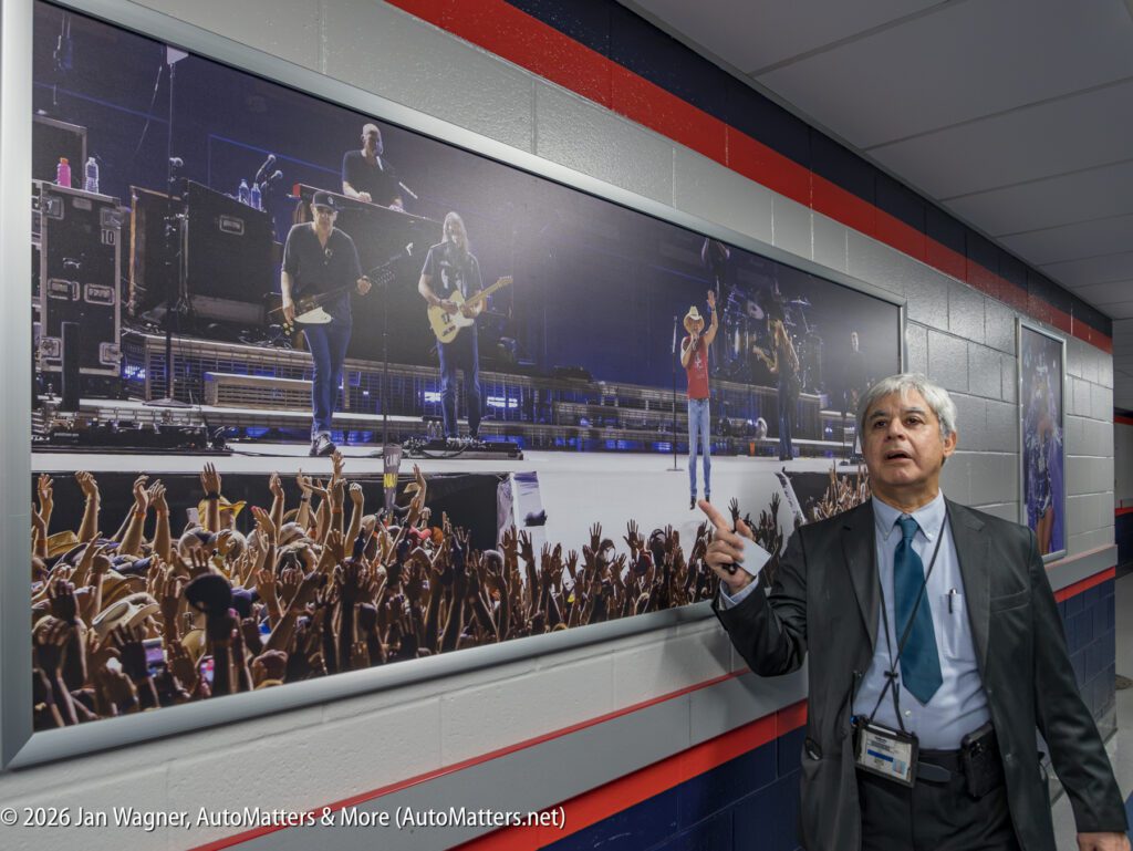A man in a suit stands in a hallway, pointing at a large framed photo of a concert crowd and band on a stage mounted on the wall.