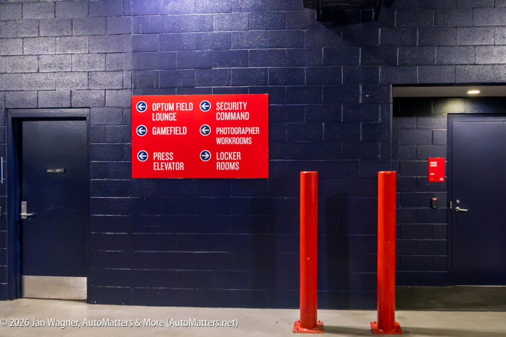 A red sign on a dark wall lists directions to the lounge, gamefield, press elevator, security, photographer workrooms, and locker rooms. Two red posts stand in front.