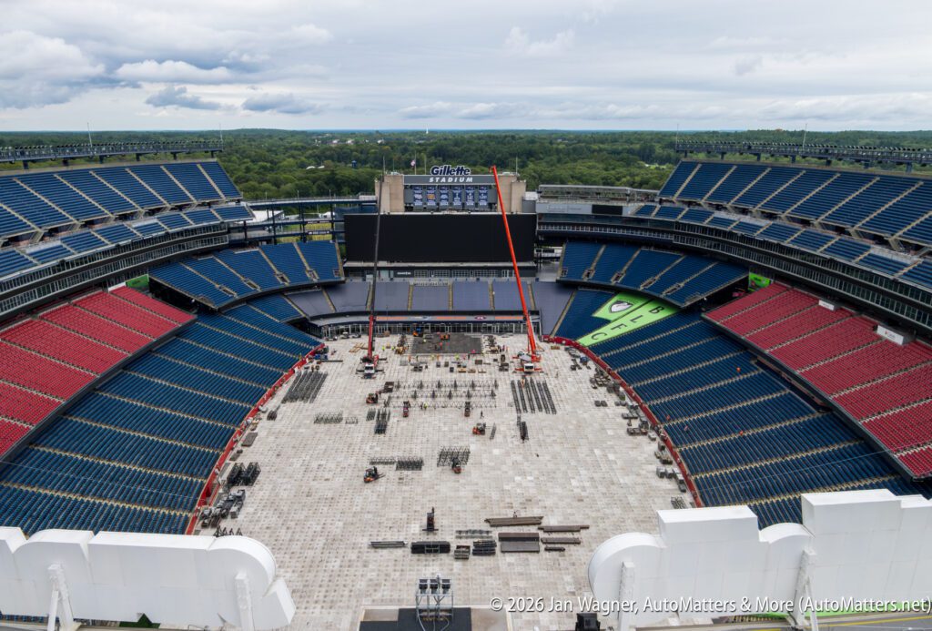 Gillette Stadium with seats mostly empty, a large stage under construction, equipment and workers on the stadium floor, and a crane beside the stage.