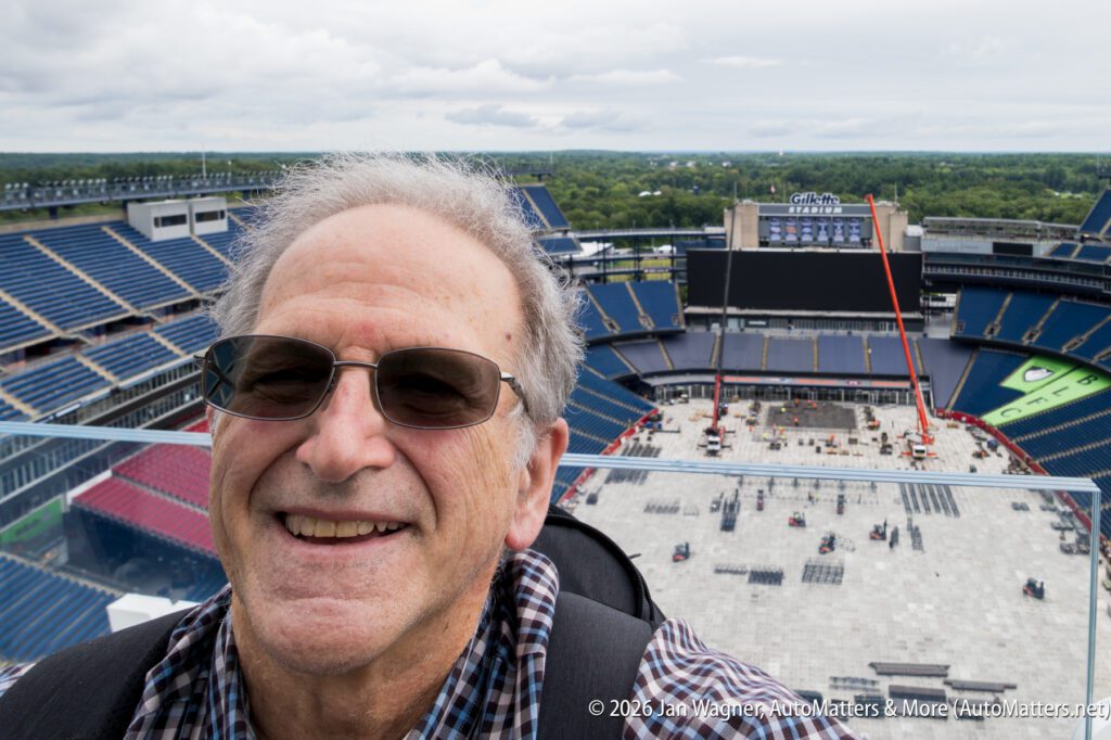 Man wearing glasses takes a selfie at a large stadium under construction, with empty seats and cranes visible in the background.
