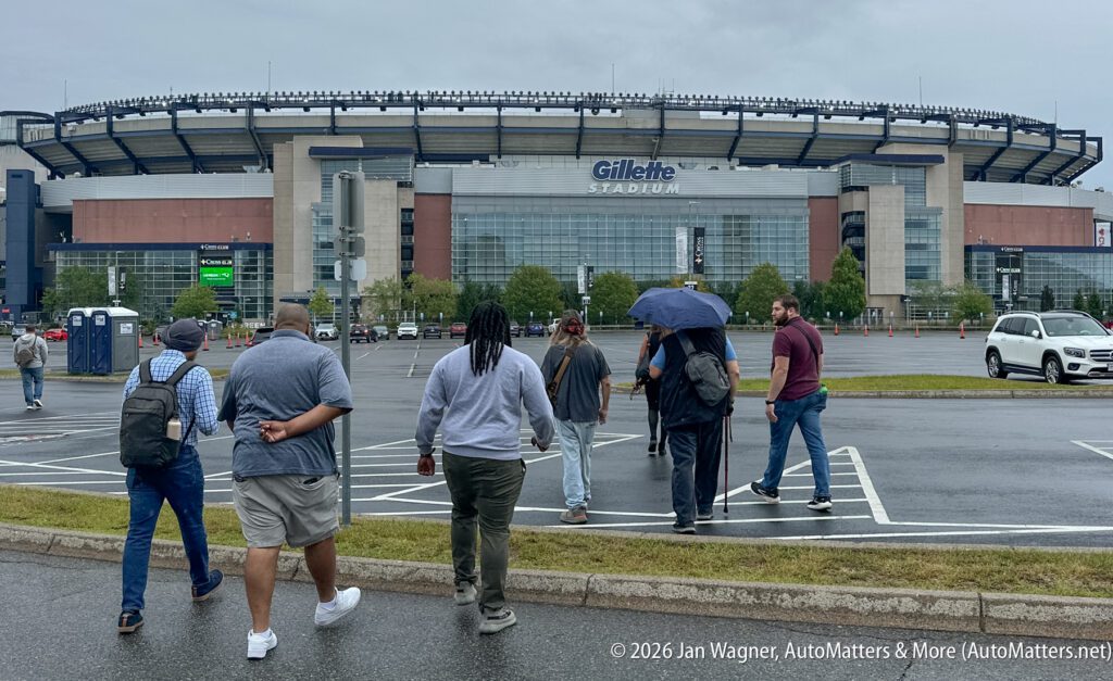 A group of people walk through a wet parking lot toward Gillette Stadium on a cloudy day.
