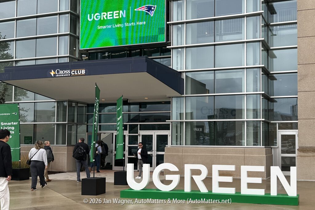 People walk into a modern glass building with large “UGREEN” signage and a digital display above the entrance, featuring the New England Patriots logo and the phrase “Smarter Living Starts Here.”.