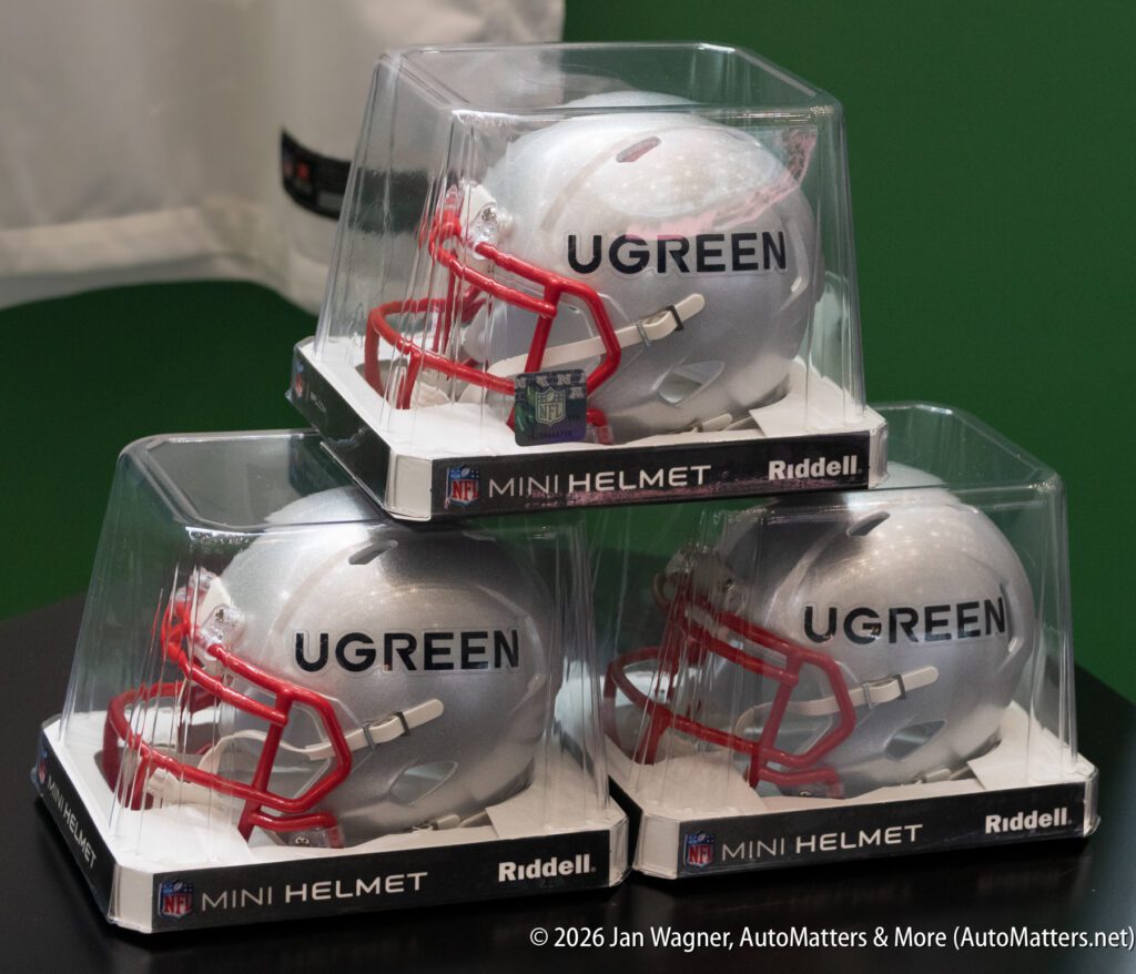 Three silver mini football helmets with red face masks, labeled “UGREEN,” are displayed in clear plastic cases on a black surface.