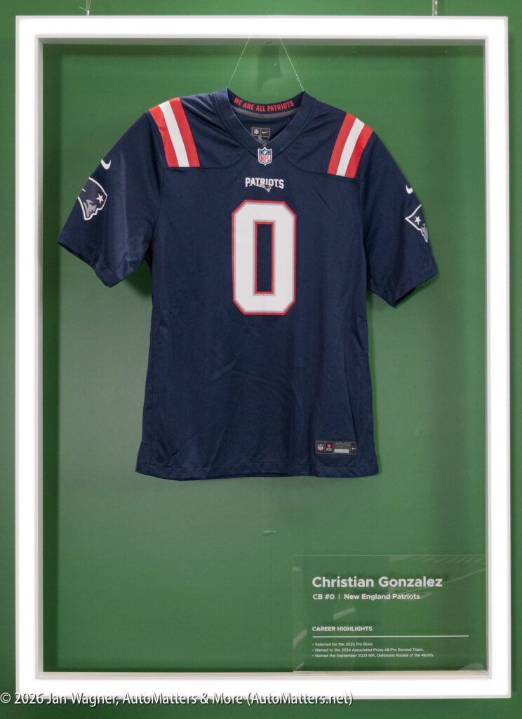 A framed New England Patriots jersey with the number 0 and “PATRIOTS” on the front, displayed next to a plaque for Christian Gonzalez.