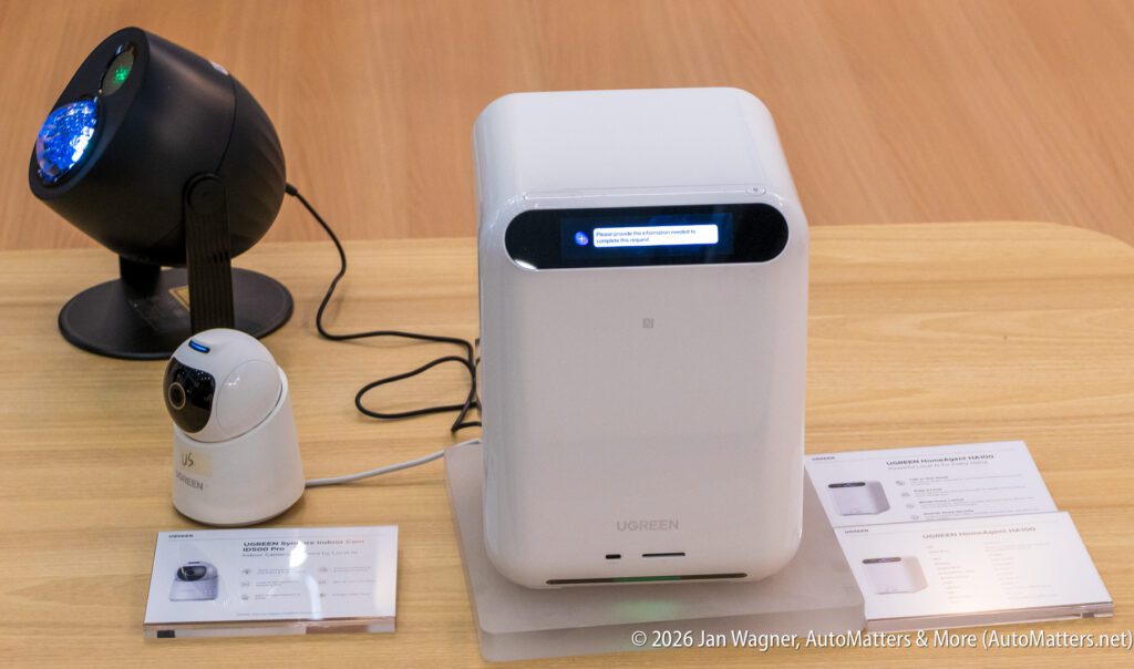 A white UGREEN charging station is displayed on a wooden table alongside a small security camera and a black device, with informational cards placed nearby.