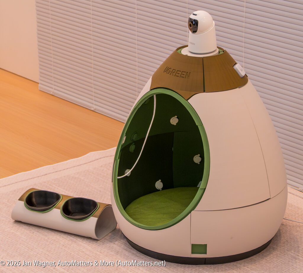 Futuristic pet house with a green interior, built-in camera on top, and a double food bowl beside it, placed on a mat indoors.
