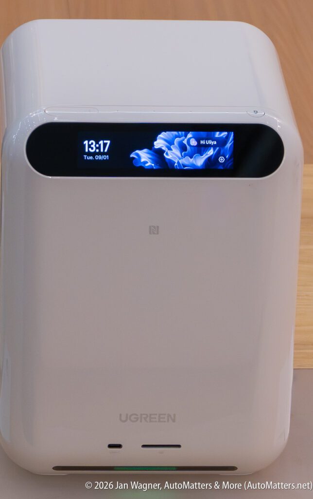 White UGREEN device with a digital screen displaying the time, date, and animated blue graphics, placed on a wooden surface.