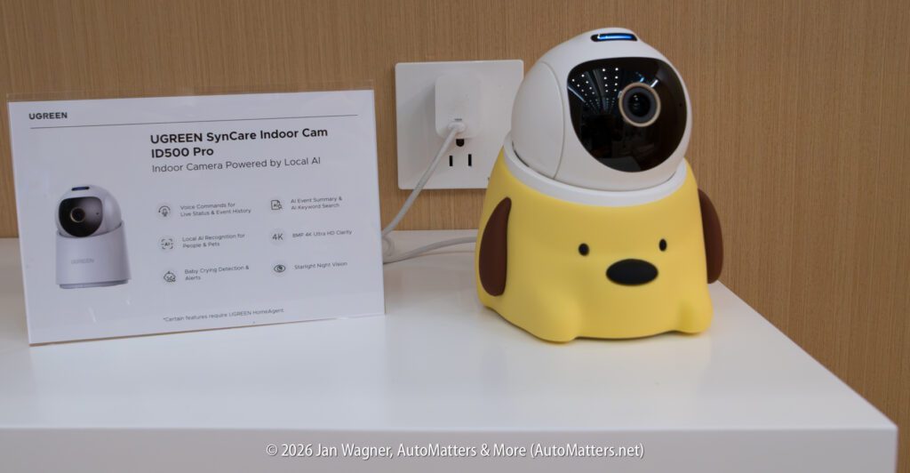 A UGREEN indoor camera in a yellow dog-shaped holder is placed on a white table next to a product info sheet and an electrical outlet.