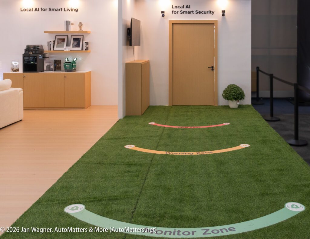 A display area with artificial grass marked as “Monitor Zone” and “Warning Zone,” adjacent to a smart living and security AI exhibit with furniture and a plant.