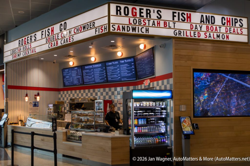 A counter-service seafood restaurant with a menu above, display case, drinks fridge, and staff member behind the counter. Signs advertise fish and chips, lobster rolls, and other seafood dishes.