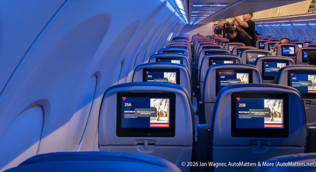 Rows of airplane seats with seatback screens displaying flight information, viewed from the aisle under blue cabin lighting.