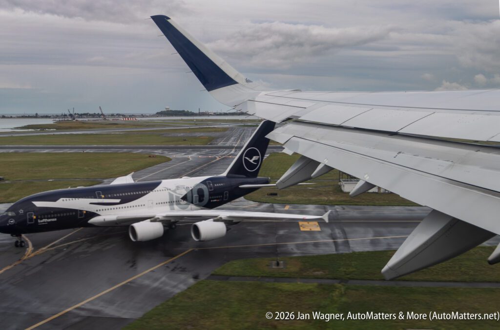 View from an airplane window showing a Lufthansa jet taxiing on a wet runway, with the wing of the photographer’s plane visible in the foreground.