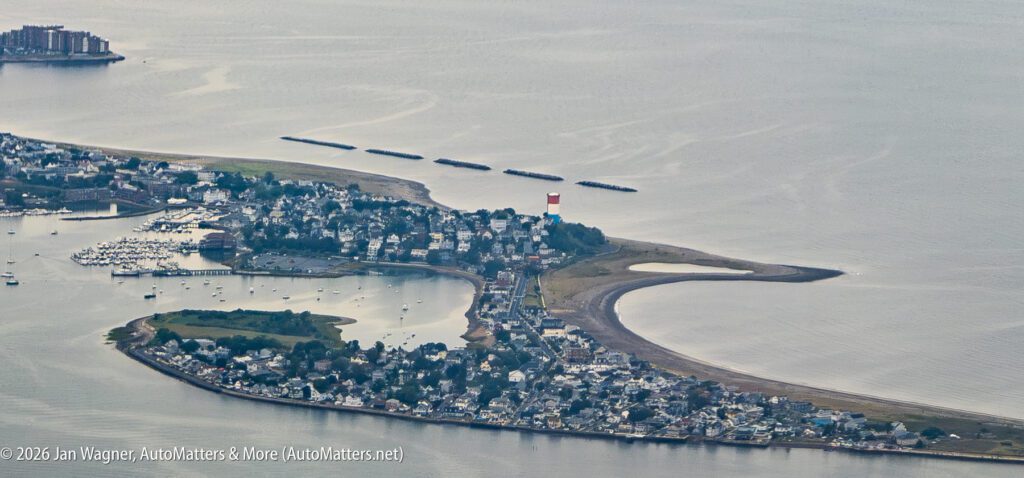 Aerial view of a coastal town with a red and white lighthouse, marina, and curving shoreline extending into the water.