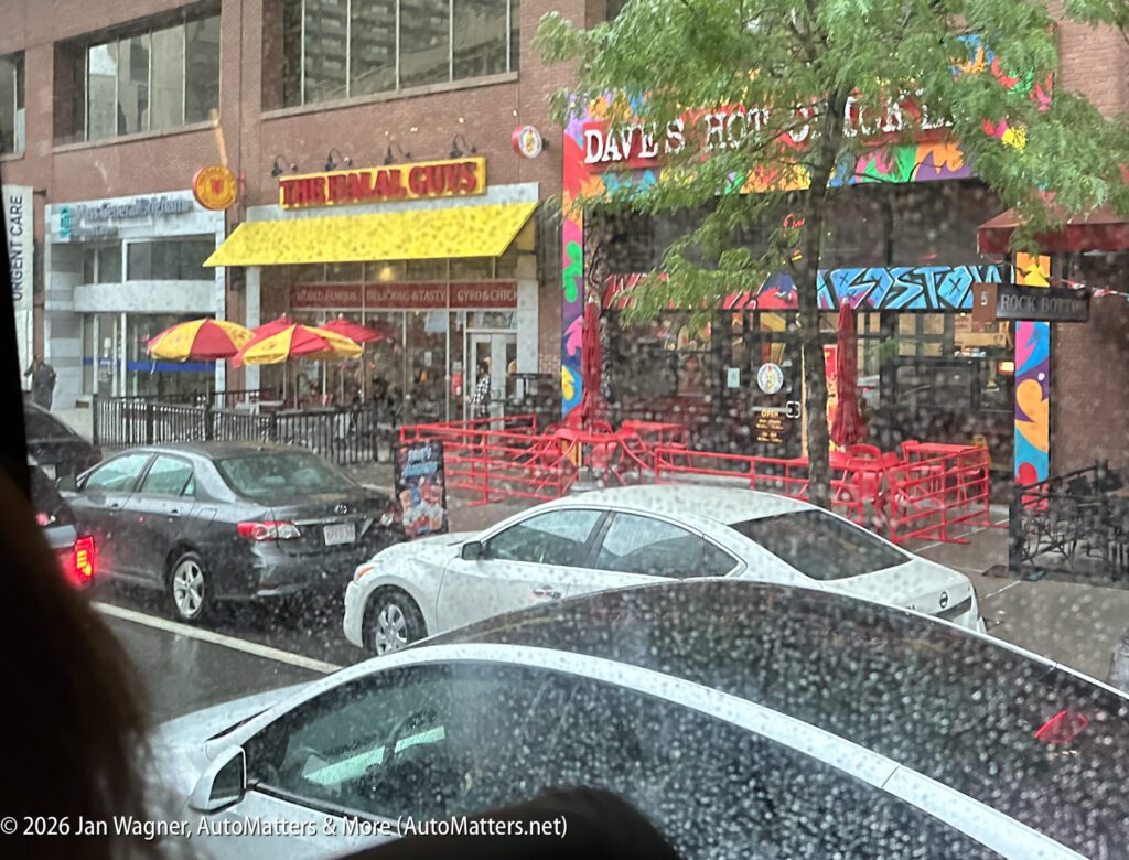 Rainy street view of parked cars in front of The Halal Guys and Dave’s Hot Chicken; red patio seating and colorful storefronts visible through a water-specked window.
