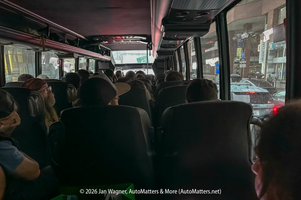 Passengers sit inside a dimly lit bus during rainy weather, looking forward through the windshield at the city street outside.