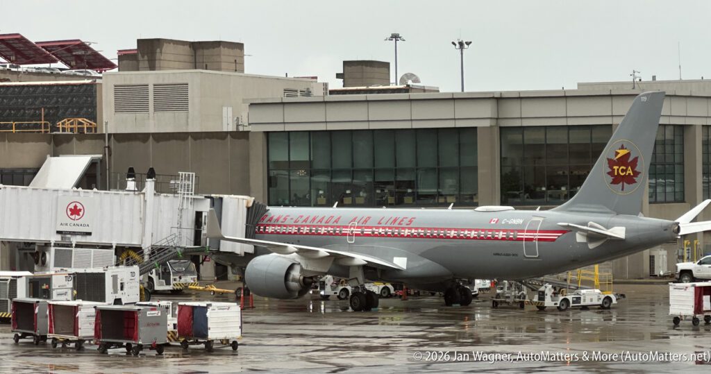 An Air Canada aircraft in retro Trans-Canada Air Lines livery is parked at a gate on a wet tarmac at an airport terminal, with ground equipment and jet bridge attached.