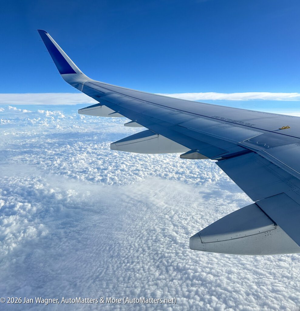 View of an airplane wing and winglet above a layer of clouds with a blue sky in the background, taken from a passenger window during flight.