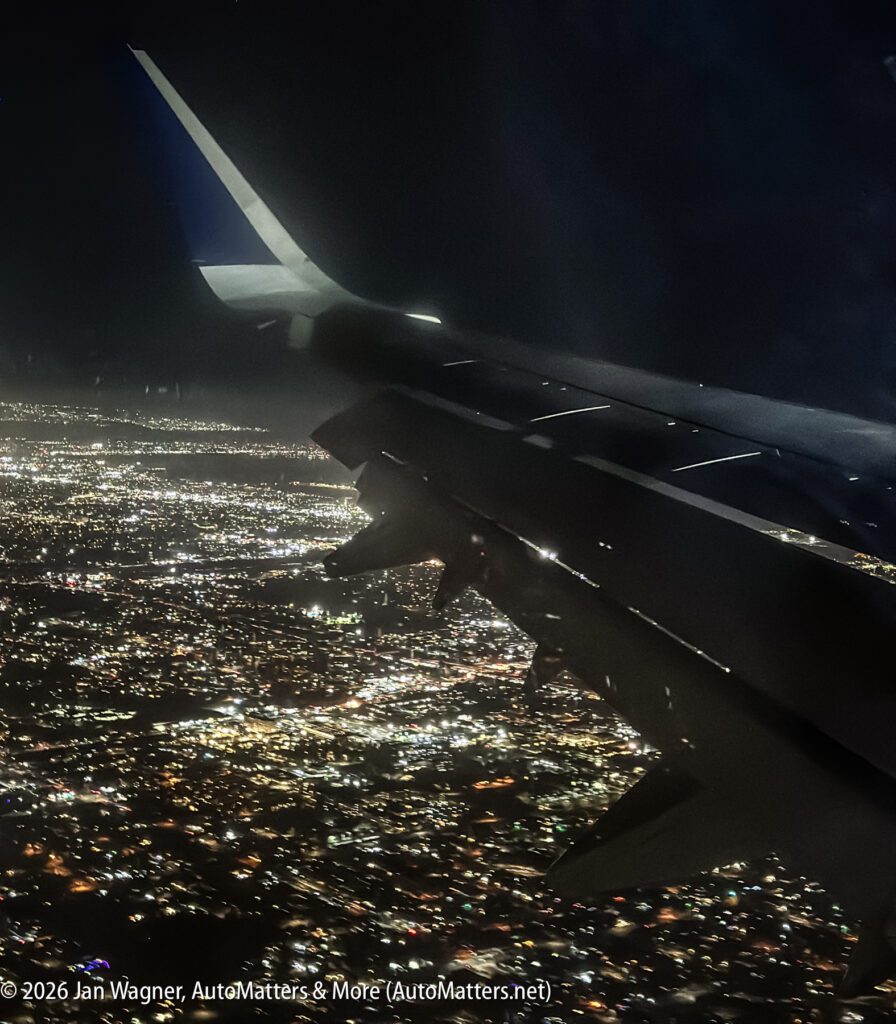 Night aerial view of city lights seen through an airplane window, with the plane’s wing and engine visible in the foreground.