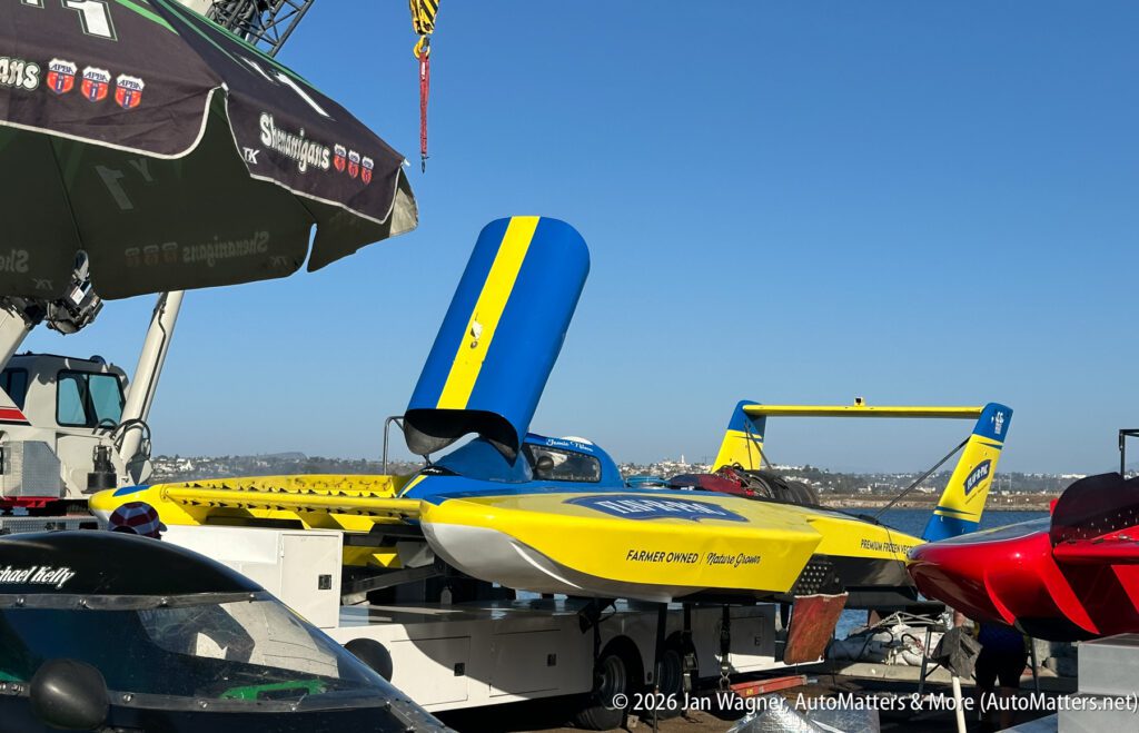 A yellow and blue hydroplane race boat with its engine cowl raised, parked on a trailer near the water.