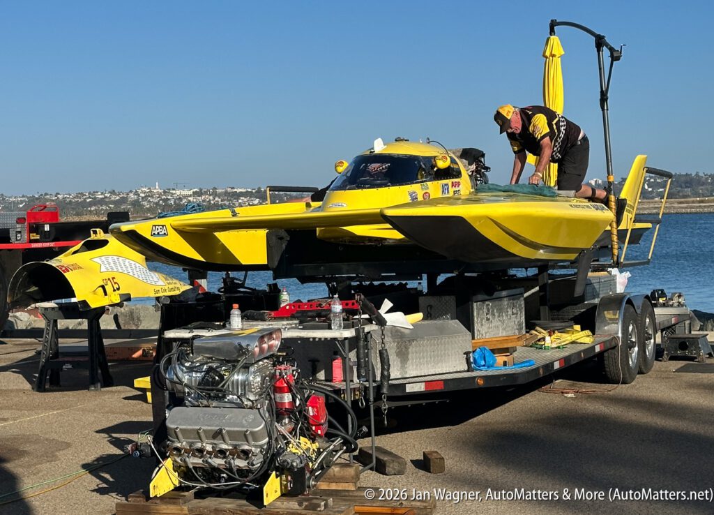A crew member works on a yellow hydroplane boat mounted on a trailer, with its engine resting on a stand nearby.