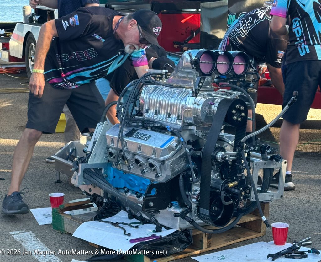A mechanic leans over a large, high-performance supercharged engine mounted on a wooden pallet.