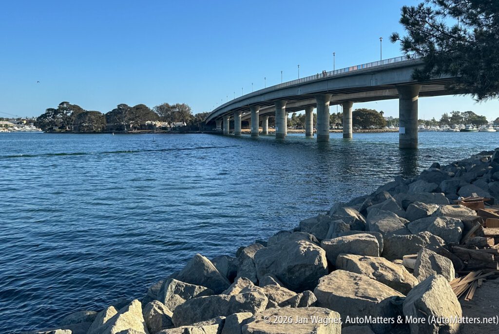 A concrete bridge spanning a wide bay, viewed from a rocky shoreline.