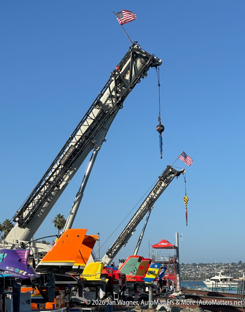 Two large cranes with American flags on their booms are positioned behind a row of parked racing boats at a marina.