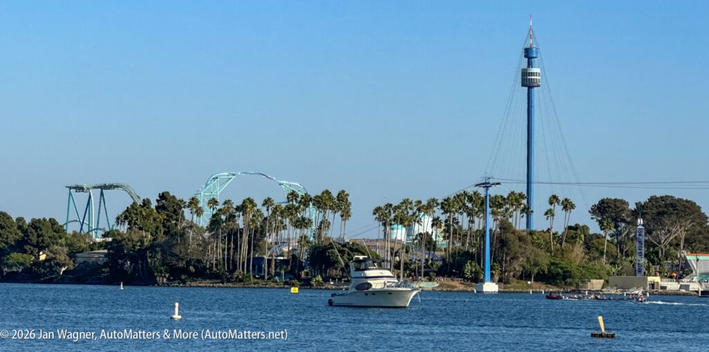 A motor yacht on a bay, with a roller coaster and an observation tower visible among trees in the background.