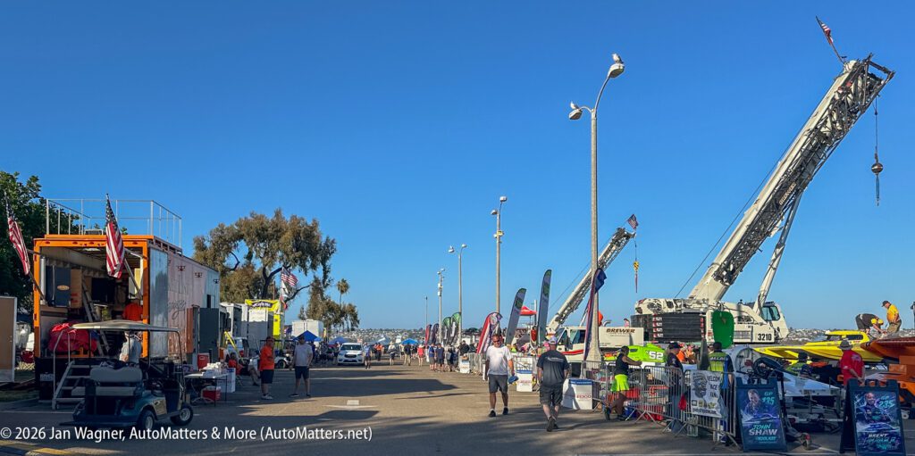 Outdoor event area with race boat trailers, large cranes, and people walking on a sunny day.