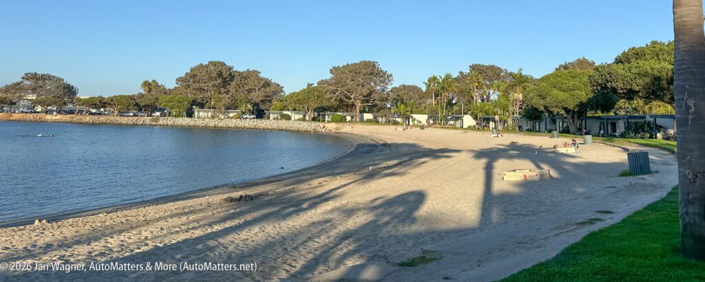 A sandy beach along a bay with small hotel buildings, trees, and a few people resting in the shade.