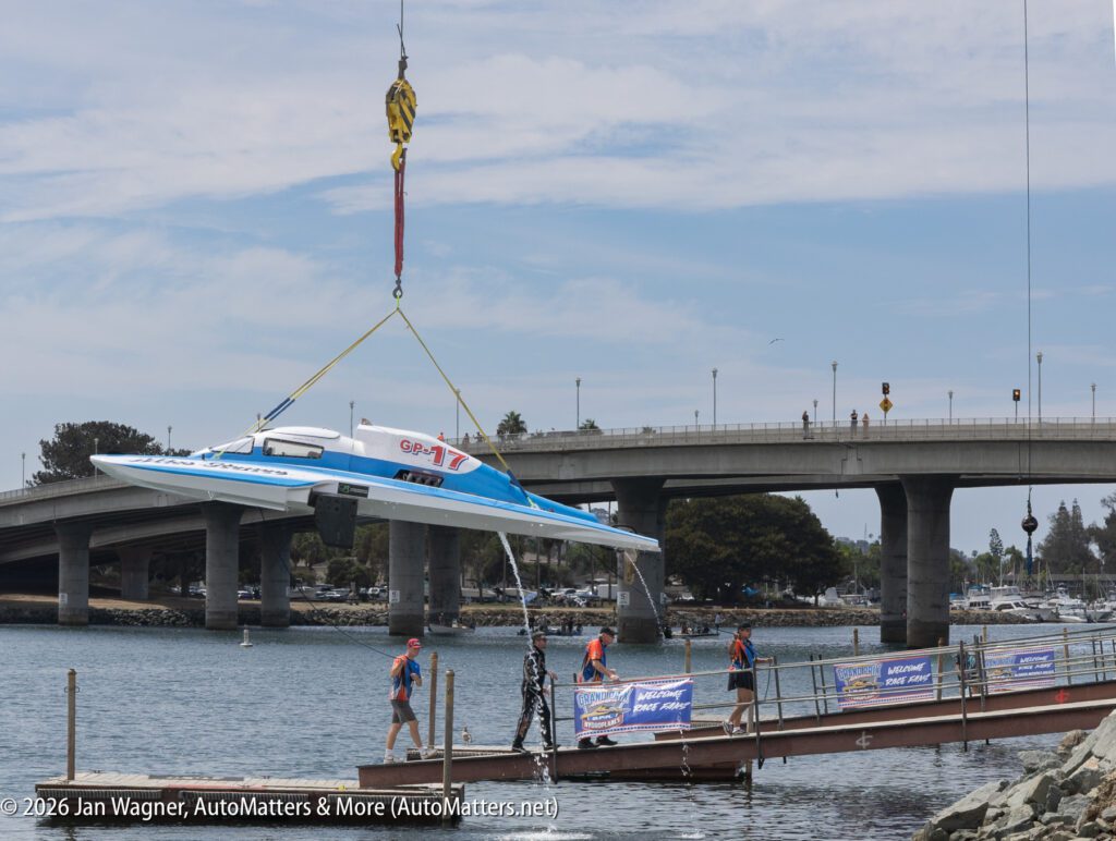 A crane lowers a blue and white racing boat marked “GP-17” toward a boat ramp where crew members stand.