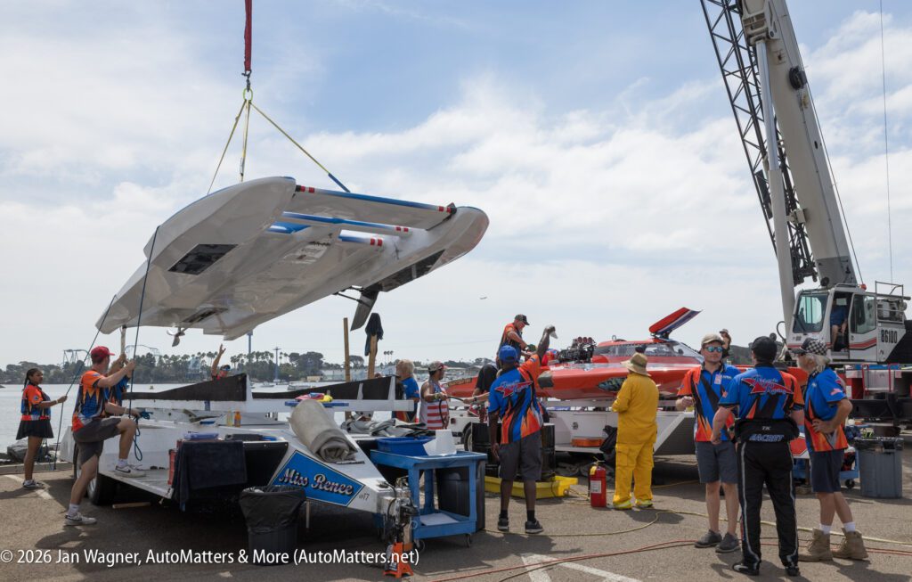 A crane lifts a large white racing boat hull above a trailer, with a crew of workers standing around it on an asphalt lot.
