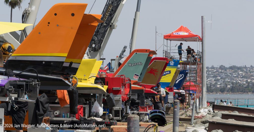 A row of hydroplane racing boats parked on trailers along a waterfront, with crew members nearby and a crane in the background.