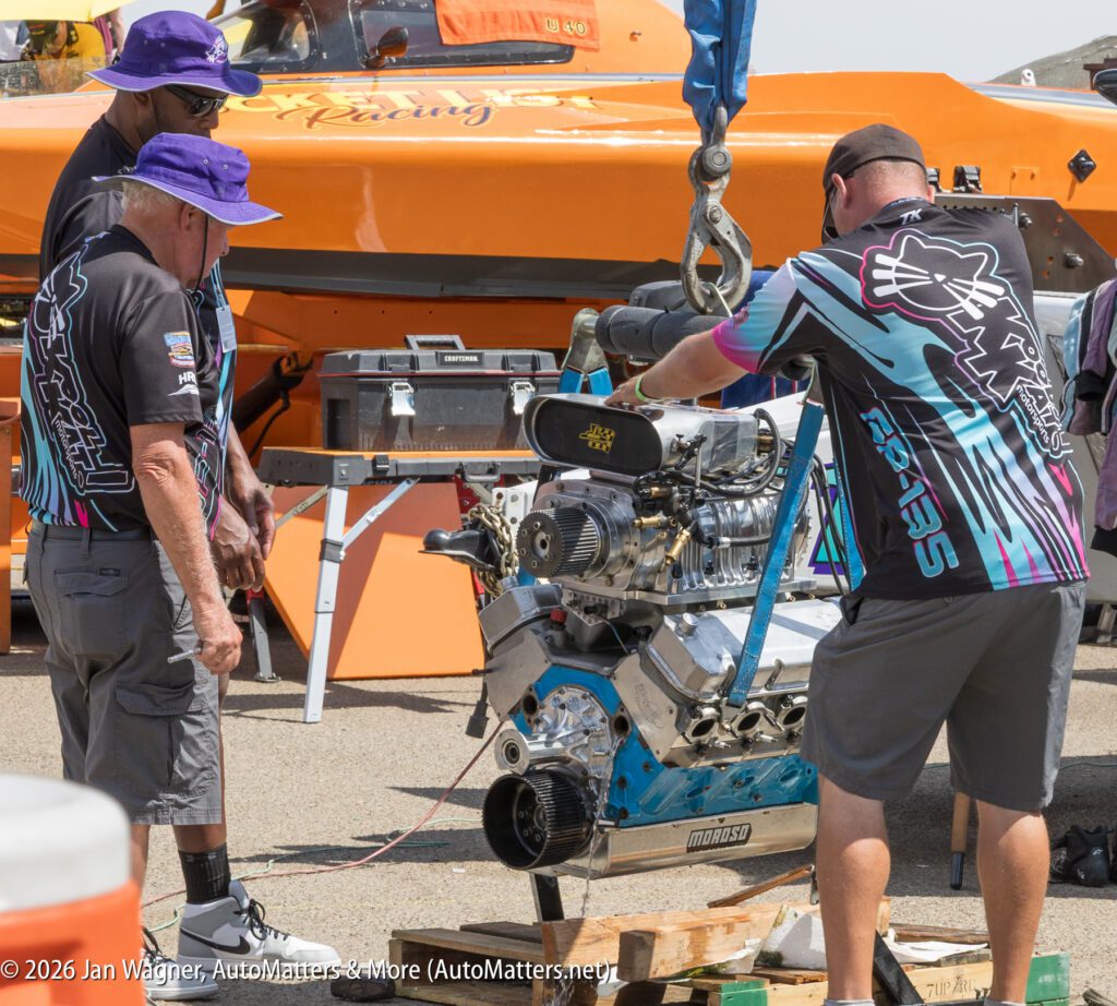 Three crew members in team uniforms work on a large exposed engine suspended by a hoist in front of an orange racing boat.