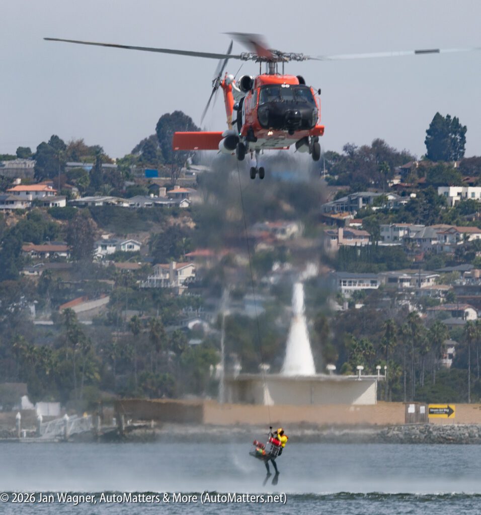 A U.S. Coast Guard helicopter hoists a rescue swimmer from the water near a shoreline.