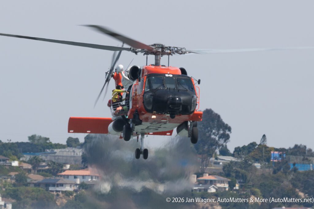 U.S. Coast Guard helicopter in flight with an aircrew member visible in the open side door.