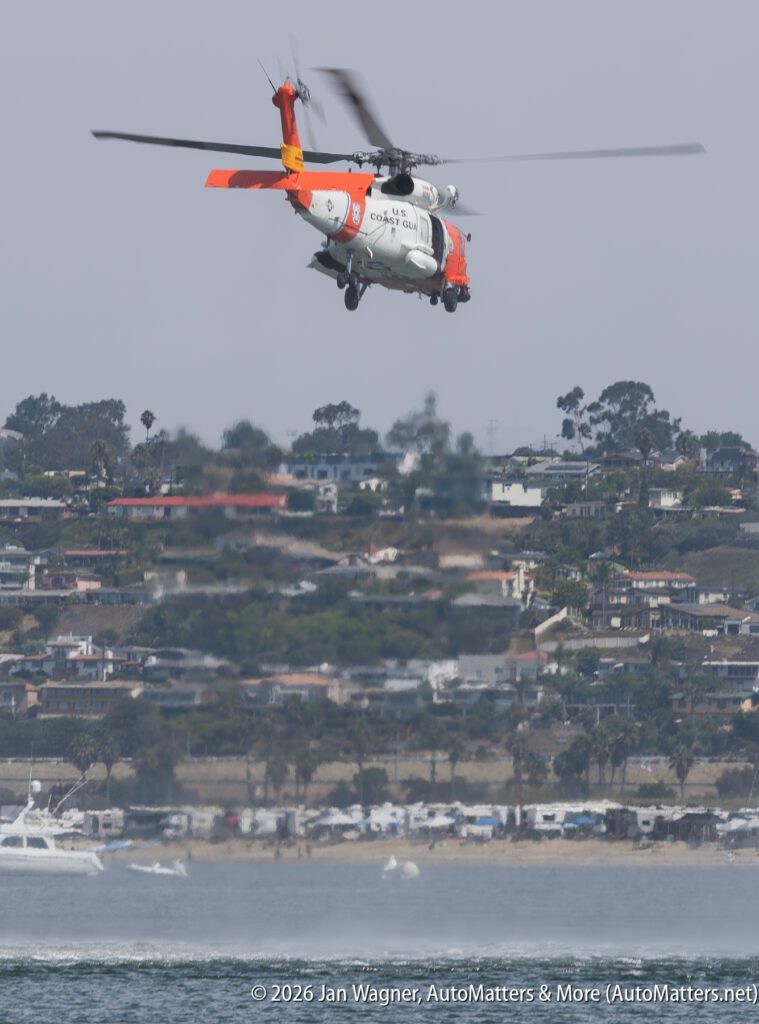 A U.S. Coast Guard helicopter hovering over the water near a beach and a residential hillside.
