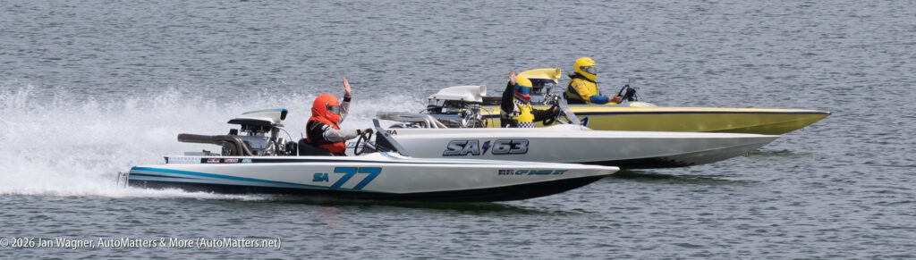 Two race boats on the water, with the drivers in orange and yellow helmets waving to the crowd.