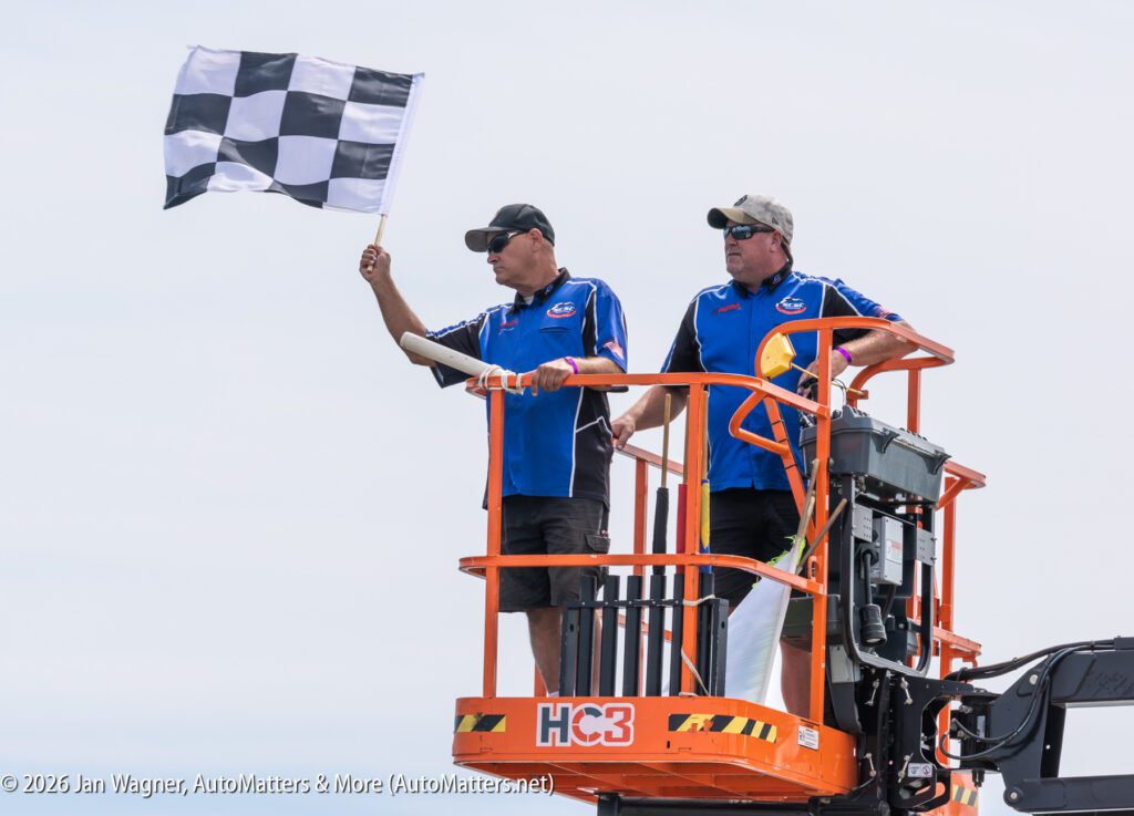 Two race officials stand in an orange aerial lift, with one holding a checkered flag.