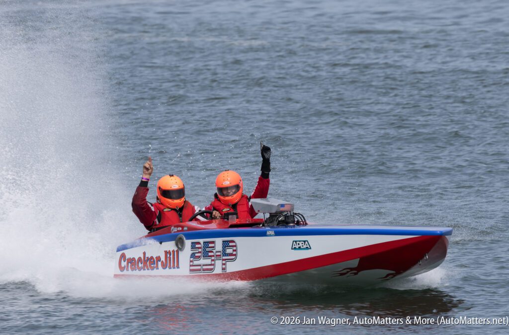 Two people in a racing boat named Cracker Jill with American flag decals on the side, both wearing orange helmets and life vests while waving to the crowd.