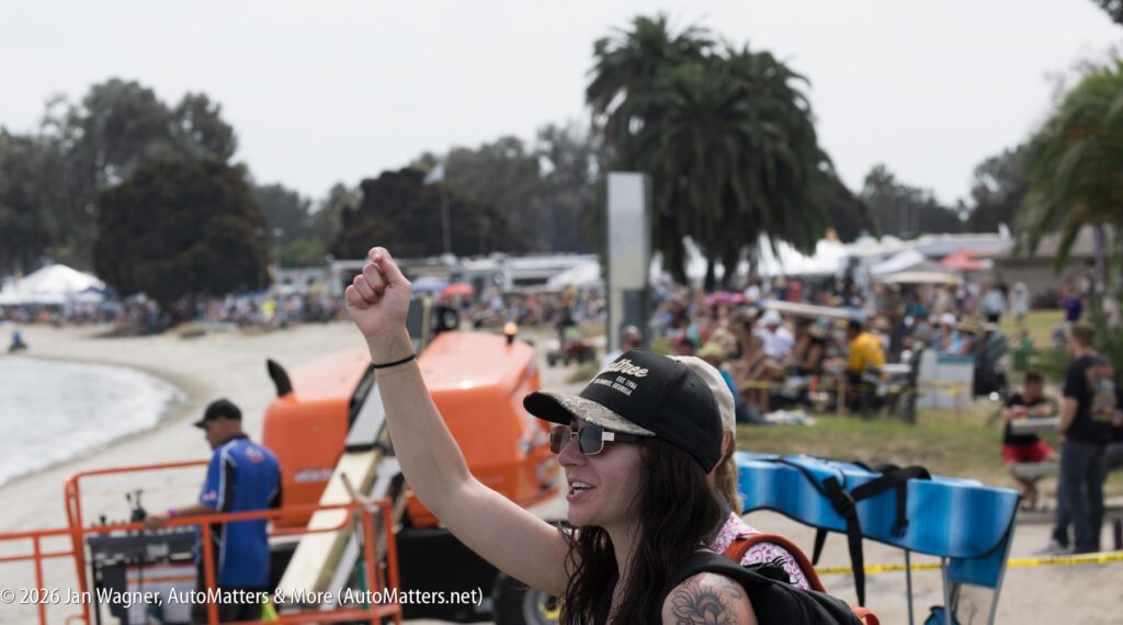 A woman wearing a baseball cap and sunglasses raises a fist in front of an outdoor crowd near a lake.